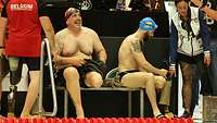Einbeinige Schwimmsportler sitzen auf einer Bank und sprechen mit rundumstehenden Personen.