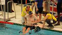 Ein gehbehinderter Schwimmsportler sitzt in einem Rollstuhl und wird mit einem Lift in ein Schwimmbecken gehoben.