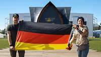 Zwei Personen halten vor dem Gebäude der Invictus games eine deutsche Flagge hoch.