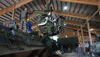 In einer Halle stehen Bundeswehrfahrzeuge. Über einem Radpanzer schwebt dessen Motor an einem Kran.