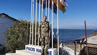 Ein Militärgeistlicher steht vor mehreren Flaggenmasten, im Hintergrund Häuser und das Meer 