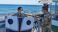 Ein Militärgeistlicher und ein Soldat bereiten eine Flagge zum Hissen vor, im Hintergrund das Meer 