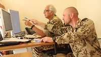 Ein Soldat hockt neben einem sitzenden Soldaten. Beide zeigen auf einen Monitor