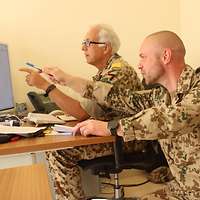 Ein Soldat hockt neben einem sitzenden Soldaten. Beide zeigen auf einen Monitor