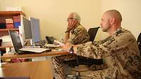 Seitenansicht auf zwei Soldaten an einem Schreibtisch, einer hockend und einer sitzend, die auf einen Monitor blicken