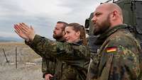 Zwei Soldaten und eine Soldatin stehen auf einem freien Feld. Die Soldatin zeigt in einer Richtung