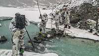 Mehrere Soldaten in Winterausrüstung überqueren einen Bach in verschneiten Bergen.
