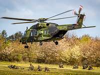 Ein NH-90 landet an Ein Hubschrauber schwebt über einem Feld. Am Boden hocken und liegen Soldaten.