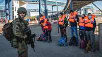 Zivilisten mit Schwimmwesten stehen nebeneinander, vor ihnen steht ein bewaffneter Soldat.