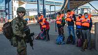 Zivilisten mit Schwimmwesten stehen nebeneinander, vor ihnen steht ein bewaffneter Soldat.