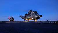 Sea King Dual Spot Nacht Zwei Hubschrauber stehen mit drehenden Rotoren auf einem dunklen Flugdeck.