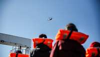 Mehrere Personen in orangefarbener Rettungsweste blicken nach oben auf einen Hubschrauber in einem blauen Himmel.