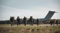 Auf einem Flugplatz steht ein großes, graues Flugzeug, davor laufen mehrere Soldaten.
