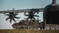 Auf einem Flugplatz steht ein großes, graues Flugzeug, davor zwei grüne kleine Panzer.