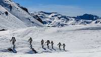 Soldaten in Schneetarn laufen hintereinander auf Skiern durch eine verschneite Gebirgslandschaft.