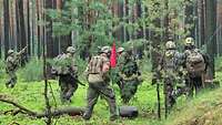 Soldaten laufen durch den Wald. Ein Soldat trägt einen roten Wimpel und eine rote Armbinde.
