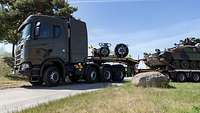 Ein mit einem Panzer beladener Lkw fährt in eine Kurve auf einer schmalen asphaltierten Straße.