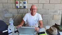A man wearing glasses and a white T-shirt is sitting in front of a laptop