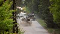 Zwei Kampfpanzer fahren hintereinander versetzt auf einer asphaltierten Waldstraße.