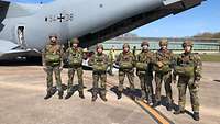 Sieben Soldaten mit angelegten Fallschirmen stehen vor einem grauen Flugzeug.