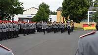 Eine Musikkapelle marschiert auf dem Appellplatz ein, dahinter die Truppenfahne und der Ehrenzug.