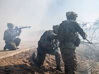 Soldaten während einer Gefechtssimulation Ein Soldat schneidet mit einem Bolzenschneider einen Stacheldraht durch. Im Hintergrund knied ein Soldat mit Waffe im Anschlag.