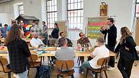 Workshop Ein Mann steht neben einem Flipchart und spricht zu einer Gruppe von Frauen und Männern, die vor ihm an einem Tisch sitzen.