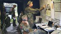Mehrere Soldaten arbeiten an Laptops in einem Zelt. Im Vordergrund steht einer vor einer Karte und ein anderer telefoniert.