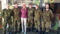 Fünf Soldaten und eine Soldatin stehen vor einer Plakatwand 