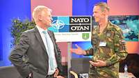 Ein Zivilist im grauen Anzug und ein Soldat stehen sich gegenüber und sprechen miteinander.