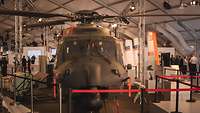 In einer Messehalle steht ein militärgrüner Hubschrauber, davor ein rotes Absperrband.