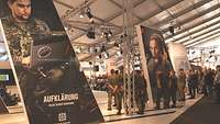 Die Bundeswehr auf der ILA22 In einer Messehalle stehen große Aufsteller mit Fotos von Soldaten. Darum herum stehen Besucher.