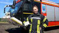 Eine junge Frau mit Zopf in schwarz-gelber Feuerwehr-Uniform steht vor einem großen Löschfahrzeug