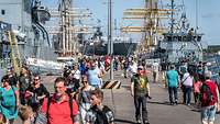 Blick entlang einer breiten Hafenmole, rechts und links liegen graue Kriegsschiffe, in der Mitte gehen viele Menschen.