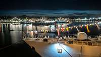 Mehrere kleine Kriegsschiffe in einem Hafen bei Nacht, sie haben Lichterketten und bunte Flaggen über ihren Aufbauten.