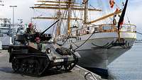 Ein kleiner Panzer steh auf einer Hafenpier vor einem dreimastigen Segelschiff.