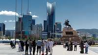 Mehrere Menschen stehen vor hohen Gebäuden und einer Statue auf einem Platz in Ulaanbaatar.