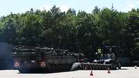 Ein Bergepanzer steht vor einem Schwerlasttransporter. Soldaten geben dem Fahrer des Bergepanzers Handzeichen.