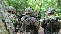 Eine Gruppe Soldaten im Gefechtsanzug steht in einem Wald im Halbkreis. In der Mitte steht ein Durchführender und spricht.