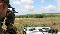 Auf einer Anhöhe liegt ein Soldat und zielt mit dem Visier seines Kompasses auf etwas. Neben ihm liegt eine Landkarte.