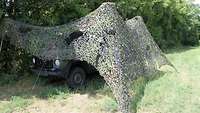 ITBtl 282 auf dem Truppenübungsplatz Ein militärisches Fahrzeug steht gut versteckt unter einem Tarnnetz an einer Waldkante.