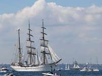 Segelschulschiff Gorchf Fock Windjammerparade Ein weißes, dreimastiges Segelschiff in See umgeben von mehreren kleinen, weißen Sportbooten.