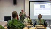 One soldier stands in front of a screen and lectures. Three others sit at individual tables and listen.