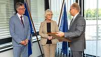 Ministerin Lambrecht steht zwischen zwei Männern in Anzügen und bekommet ein gerahmtes Dokument überreicht