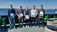 Fünf junge Männer stehen nebeneinander auf dem Deck eines Schiffes