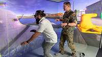 Ein Mann mit 3D-Brille und Joystick vor einem U-Boot. Neben ihm steht ein Soldat.