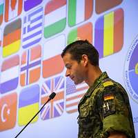 Der Soldat steht an einem Mikrofon. Hinter ihm werden verschiedene Flaggen der CWIX projiziert. 