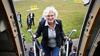 Eine Frau steigt über eine Leiter mit Handgriff in ein Flugzeug; im Hintergrund eine Grasfläche außen vor dem Flugzeug.