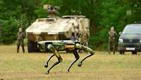Ein Roboterhund läuft über eine Wiese. Im Hintergrund stehen Soldaten und Militärfahrzeuge.