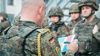 Ein Soldat mit gelber Kordel steht vor einer Truppe angetretener Soldatinnen und Soldaten und hält einen Zettel in der Hand